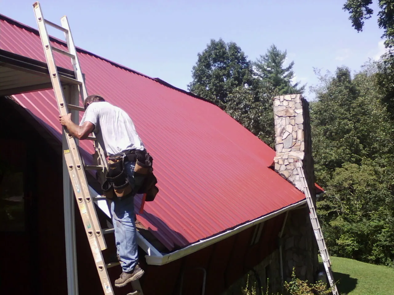 Roofer on a ladder beside a red metal roof and stone chimney — Soffit & Fascia Repair by Roofing Brothers in Bellbrook
